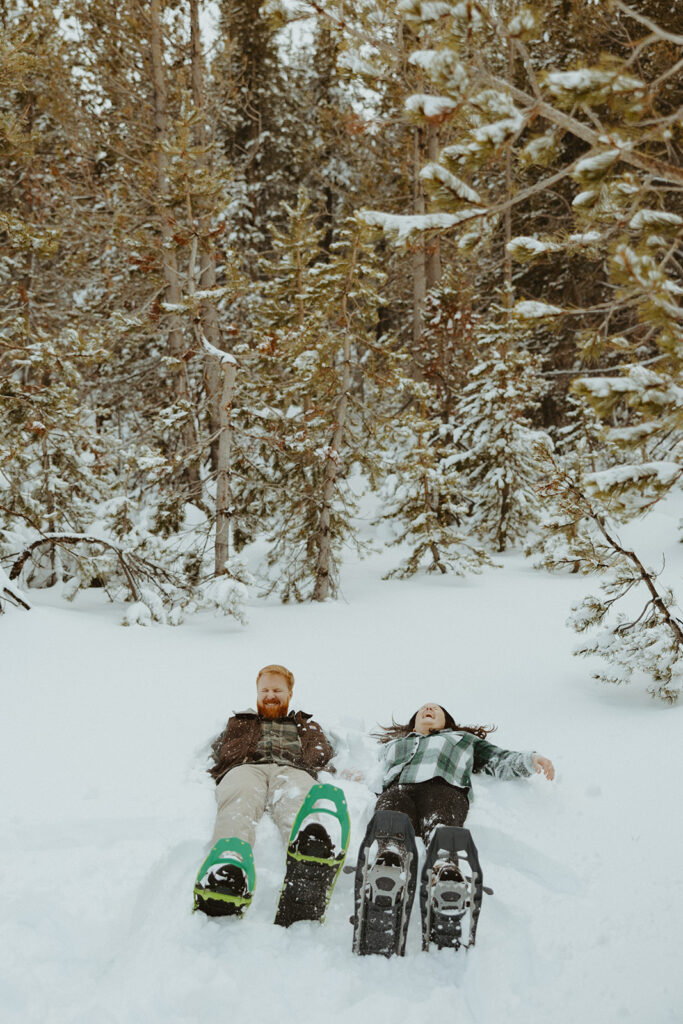 couple laying down in the snow with snow covered trees behind them