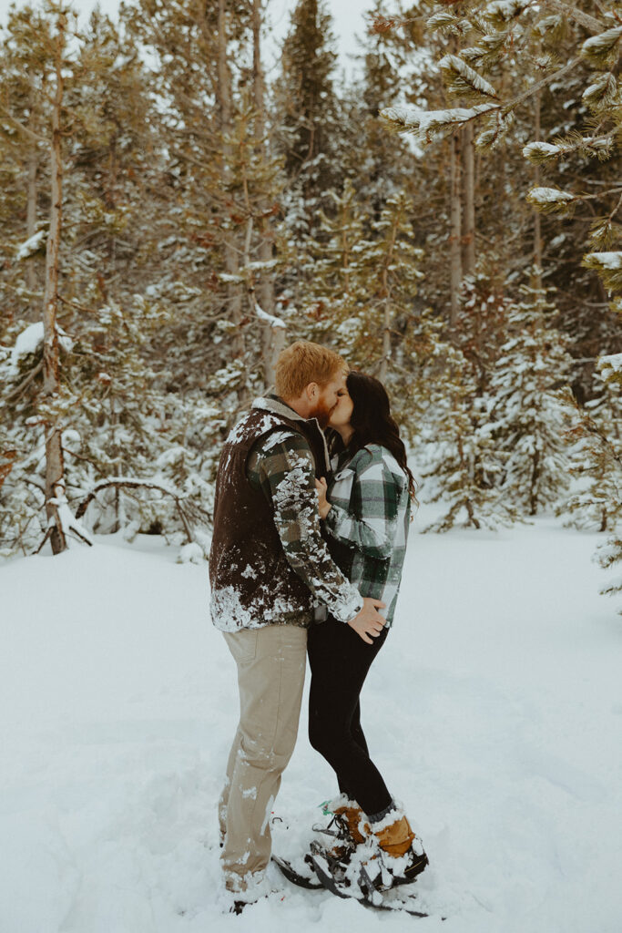 husband and wife kissing in the snow with pine trees behind them