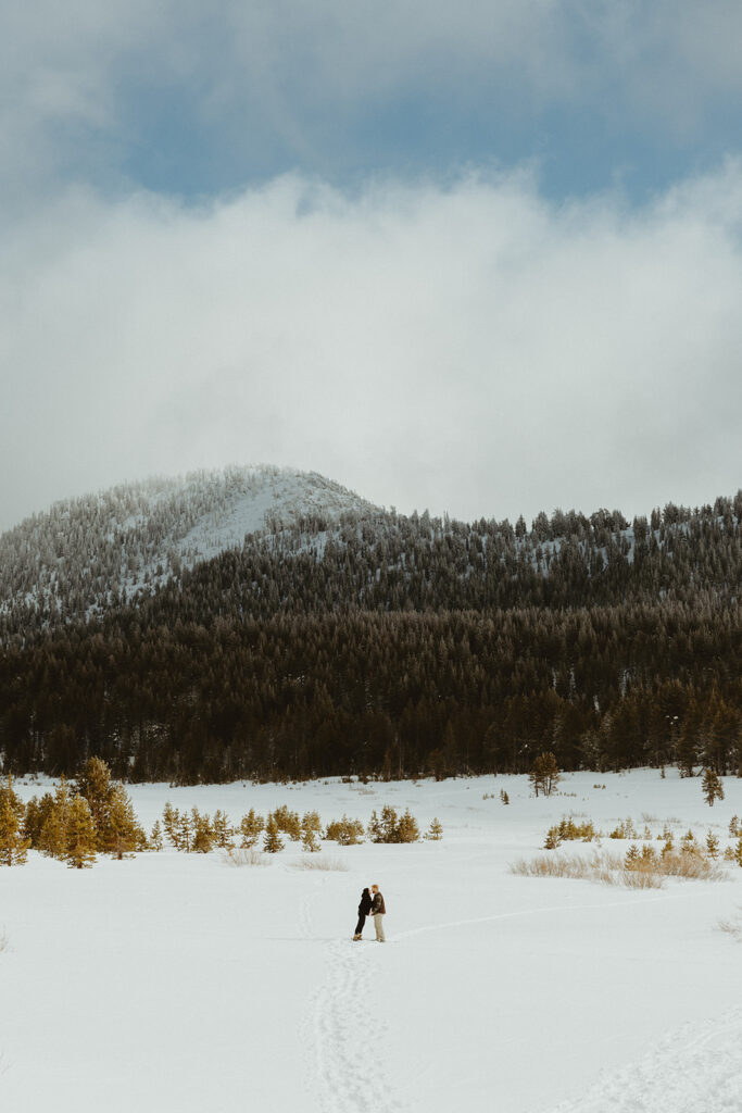 couple kissing in the snow from afar with a mountain behind them