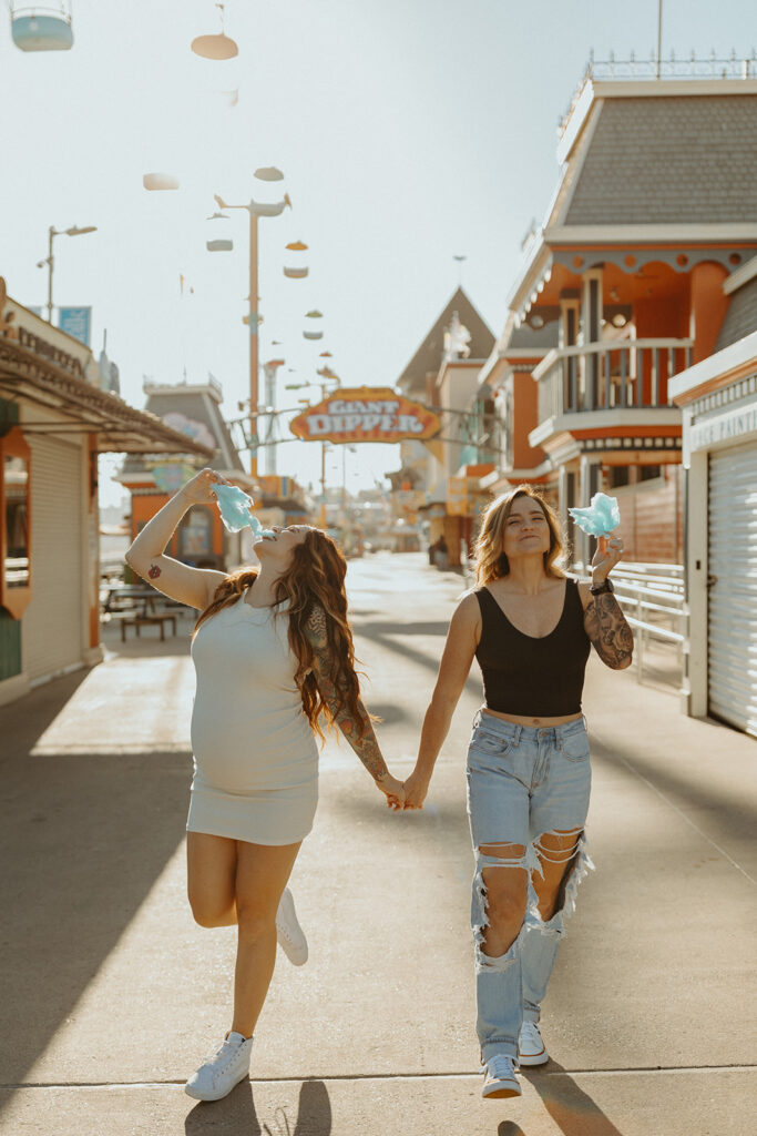 Couple holding hands and eating blue cotton candy while walking down the boardwalk for their Santa Cruz maternity photoshoot