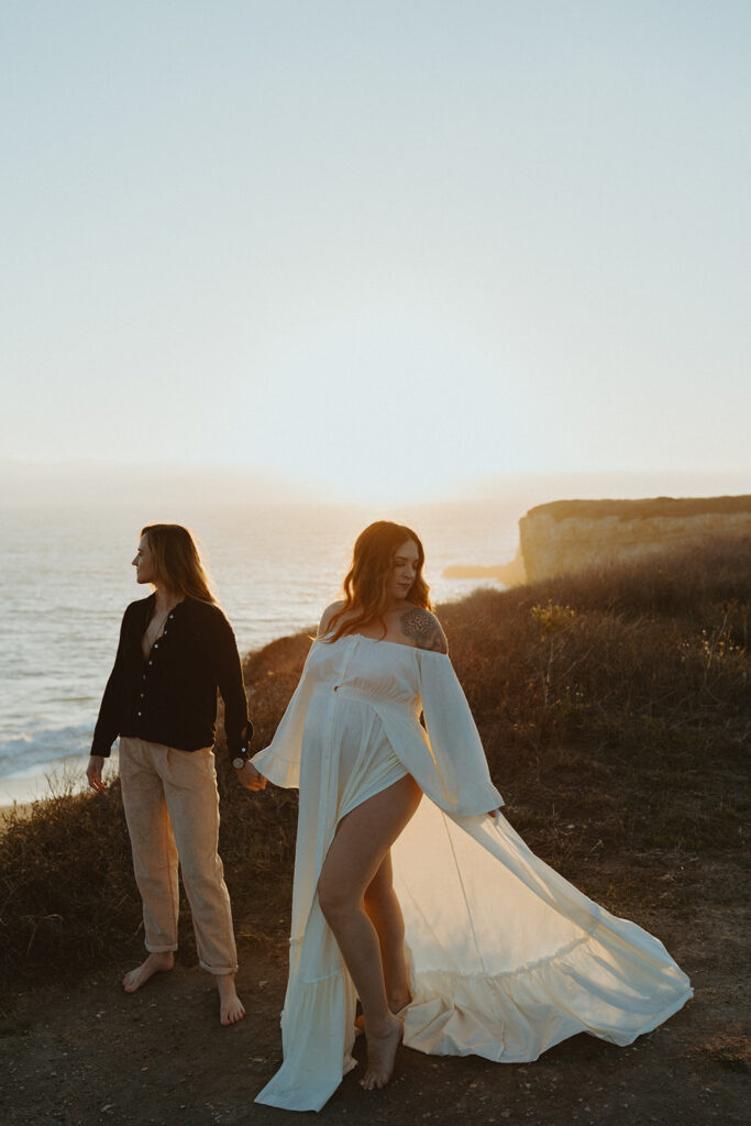 santa cruz maternity photoshoot of Roni and Cassi looking opposite directions and holding hands on a cliffside at sunset