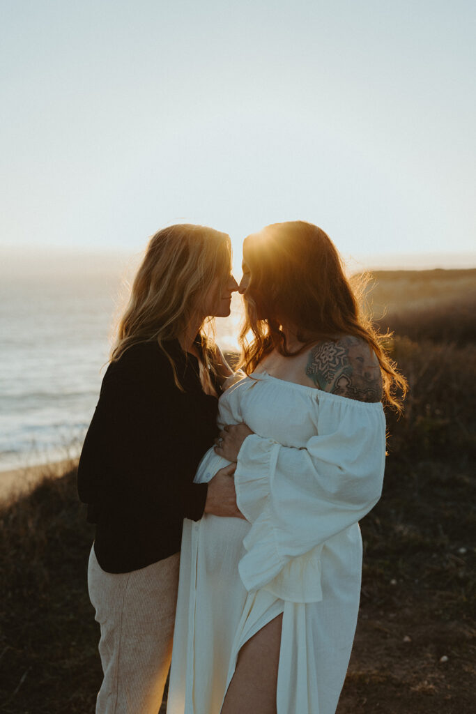 Roni and Cassi touch noses on the cliffs on the beach and holding the baby bump at sunset