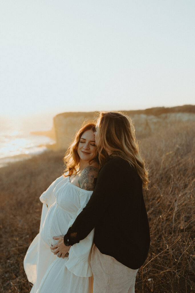 Roni holding her dress out and Cassi holding the baby bump overlooking the ocean on the cliffs