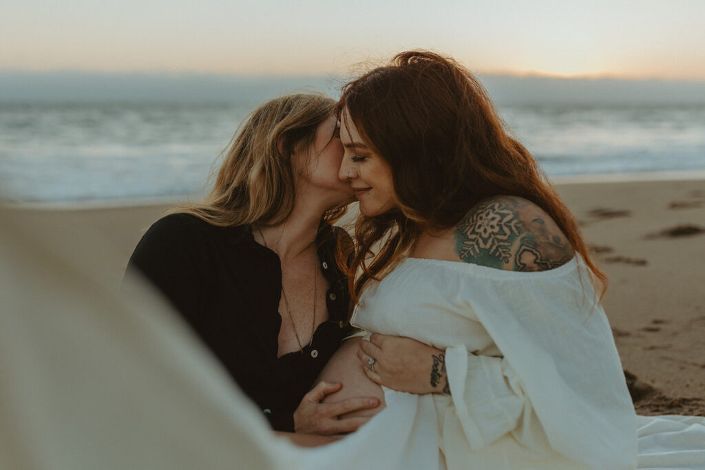 Roni and Cassi sitting in the sand holding the baby bump